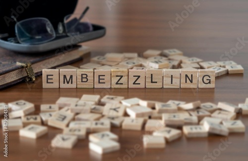 embezzling concept represented by wooden letter tiles on a wooden table with glasses and a book