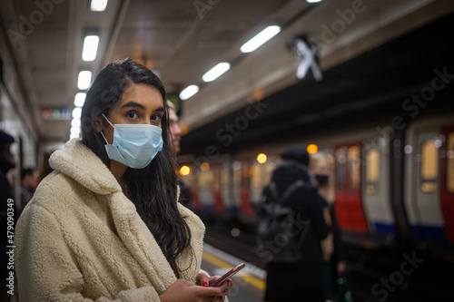 Girl waits for the subway in the London Underground, she wears a mask to use public transport and holds her phone in her hand