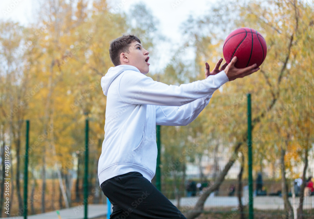 Fototapeta premium Cute teenager in white hoodie playing basketball. Young boy with red ball learning dribble and shooting on the city court. Hobby for kids, active lifestyle