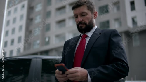 Business man texting phone in the street. A serious middle-aged businessman in luxurious clothes in the financial district of a metropolis uses a mobile phone for negotiations. photo