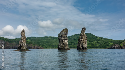 Picturesque high cliffs rise above the Pacific Ocean. Bird nests on steep slopes. The green hilly coast of Kamchatka against the blue sky. Rocks Three Brothers