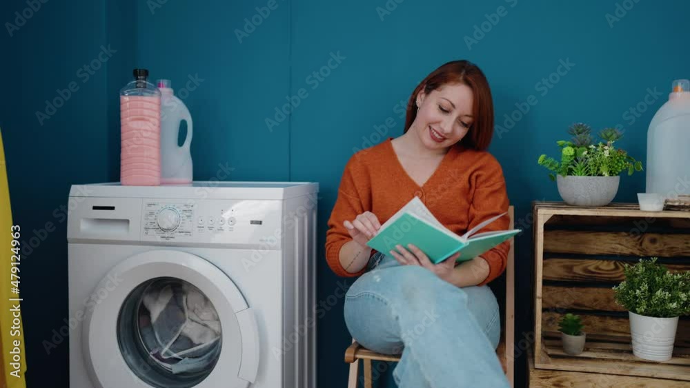 Young redhead woman reading book waiting for washing machine at laundry room