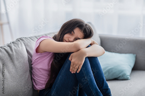 Unhappy teen girl feeling depressed or desperate, sitting on couch at home, suffering from problem or teenage depression photo