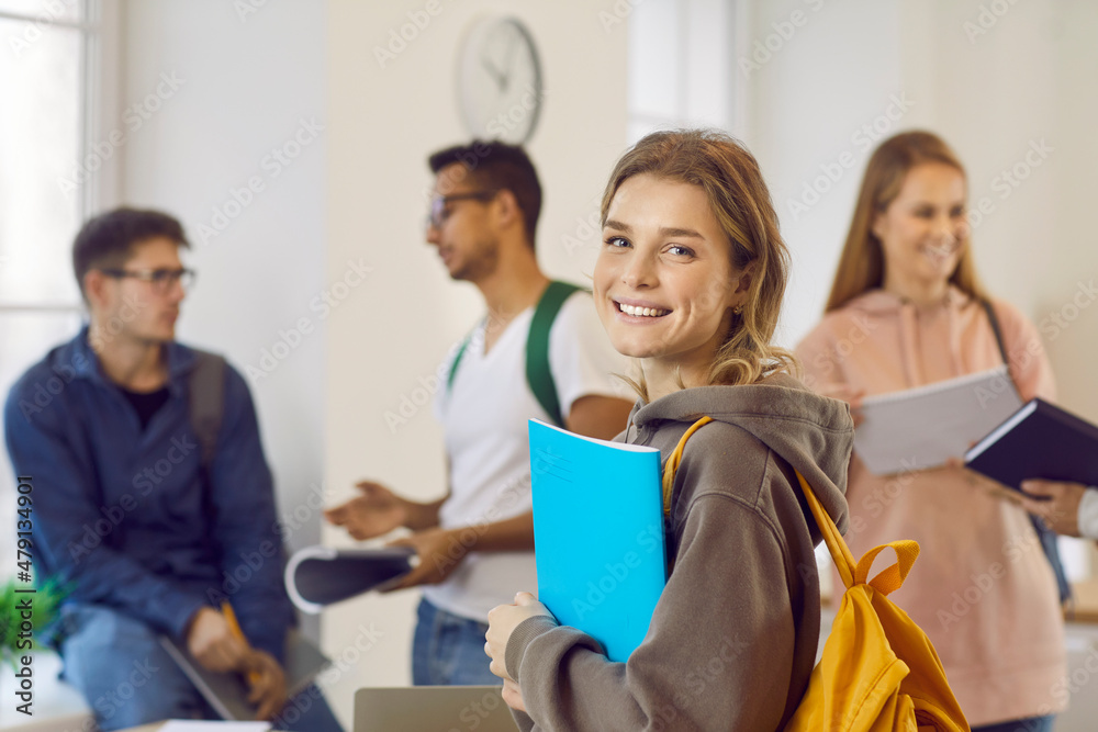 Happy beautiful female university student with book and backpack. Portrait of pretty young girl with candid cheerful face expression smiling at camera while standing in classroom with her classmates Happy beautiful female university student with book and backpack. Portrait of pretty young girl with candid cheerful face expression smiling at camera while standing in classroom with her classmates