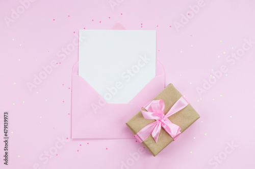 White empty card with copy space in pink envelope on pink background with gift box wrapped in craft paper © Eugenia