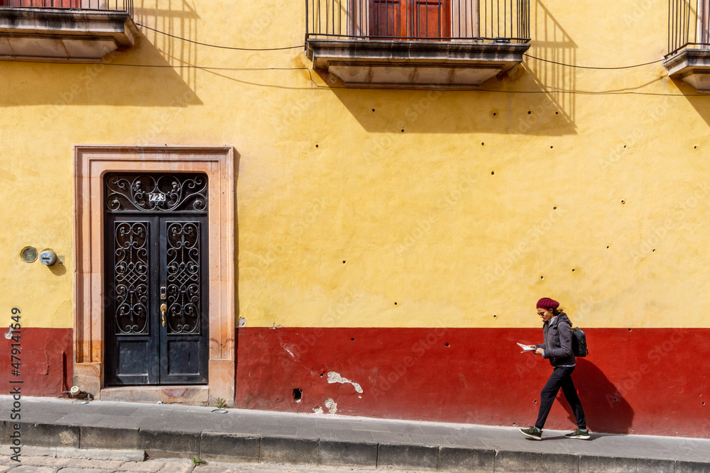 A slender woman walks in front of a colonial-style facade while she reads a paper map
