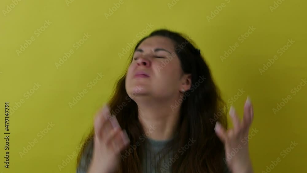 A beautiful indignant woman in a gray sweater on a yellow background nervously gesticulates with her hands. Pretty nervous woman with long hair waving her hands with negative emotion.