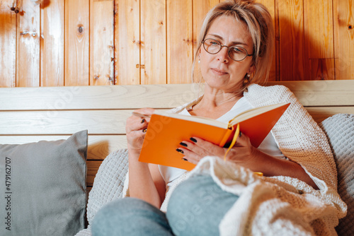 Close-up caucasian adult woman wearing glasses freelancer or student with diary sitting on bed in bedroom, indoors. Middle aged woman taking notes, reading something in notebook photo