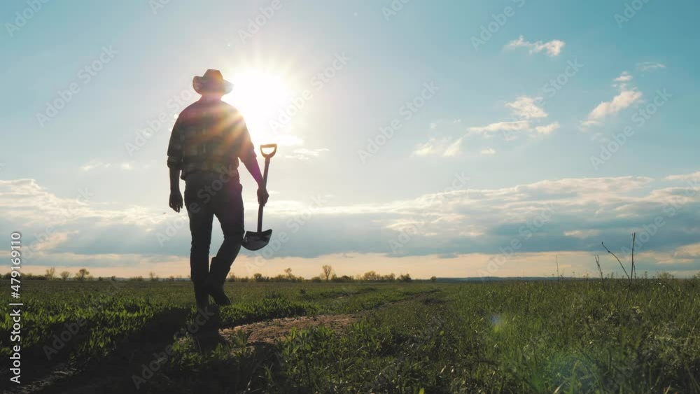 Farm agriculture concept. Farmer with a shovel in rubber boots walks across field at sunset. Silhouette man gardener walking along the road along the field. Tired man after hard work on the ground. Farm agriculture concept. Farmer with a shovel in rubber boots walks across field at sunset. Silhouette man gardener walking along the road along the field. Tired man after hard work on the ground.
