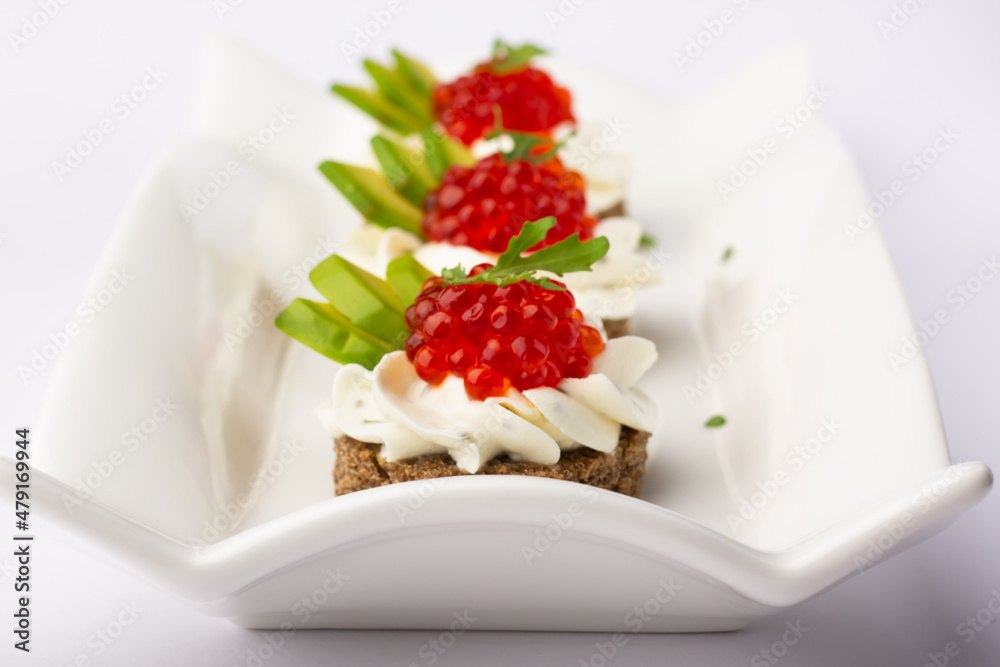 Smorrebrod. A delectable ensemble of red caviar canapés, accentuated with fresh avocado and cream, artistically placed on a uniquely shaped white dish for a modern touch. Smorrebrod. A delectable ensemble of red caviar canapés, accentuated with fresh avocado and cream, artistically placed on a uniquely shaped white dish for a modern touch.