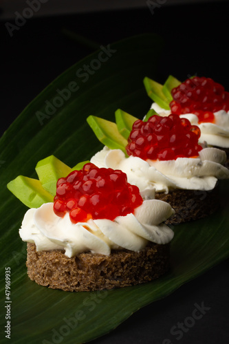 Smorrebrod. Elegant gourmet canapés with radiant red caviar atop a velvety cream, nestled on rye bread and paired with vibrant green avocado, set against a leafy backdrop.
