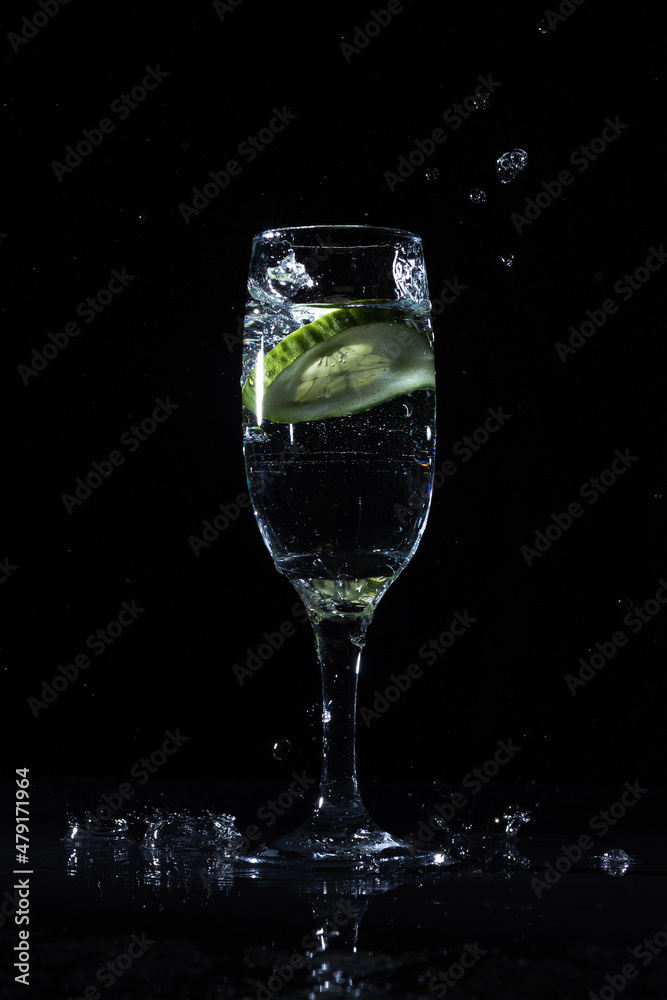 Piece of fresh cucumber splashing infused water in wineglass on black background in dark studio showing detox drink concept Piece of fresh cucumber splashing infused water in wineglass on black background in dark studio showing detox drink concept