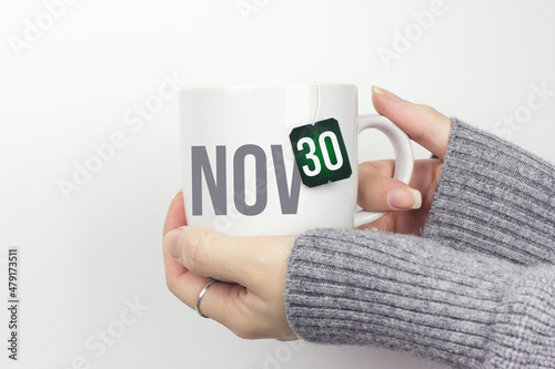 November 30th. Day 30 of month, Calendar date. Closeup of female hands in grey sweater holding cup of tea with month and calendar date on teabag label. Autumn month, day of the year concept.