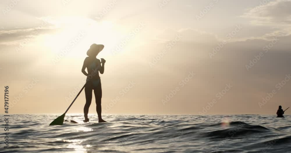 Young woman silhouette wearing bikini swimsuit and hat swims on stand-up paddle board enjoying sea and sky view at bright sunset