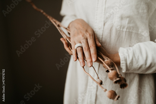 A woman is wearing a white cotton macrame ring while she is holding a dried poppy bunch. She is wearing white linen blouse. Copy space image. Sustainable fashion trendy accessory. 