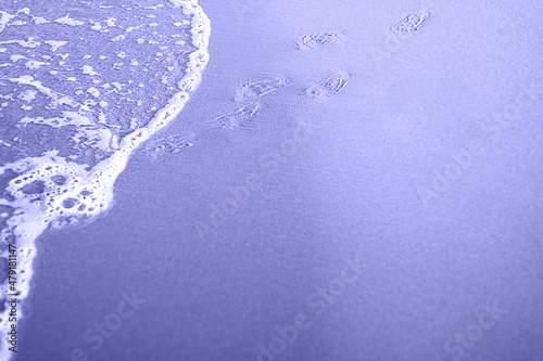 Toned with Very Peri color Kid footprints on sand at the Beach and cooming foamy wave. Smooth costline. Horizontal with copy space. Travelling and holiday concept photo