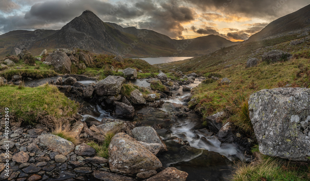 Epic moody Autumn sunset landscape image of Llyn Ogwen and Tryfan in Snowdonia National Park with stream and rocks in foreground
