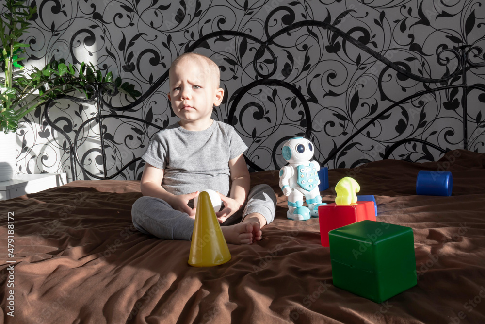 Naklejka premium A little boy is playing with a robot and various plastic toys, building a constructor on a bed in the interior of a room.