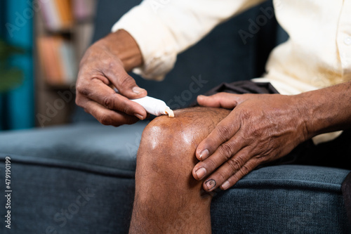 Close up of unrecognizable old man applying ointment cream for joint knee pain at home - concept of treatment or therapy for osteoarthritis and knee sprain