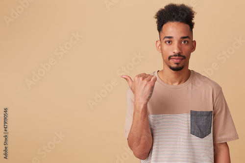 Studio portrait of young african american male points aside at copy space with excited  shocked facial expression. posing over beige background