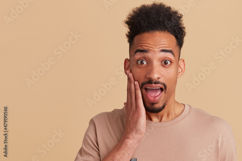 Studio portrait of young african american male looking directly into camera with. shocked, surprised facial expression, touching his cheek. posing over beige background © timtimphoto
