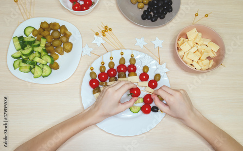 The child's hands are preparing canapes for a holiday party. The ingredients are laid out on the table: cheese, cucumbers, olives, cherry tomatoes. Step by step. photo