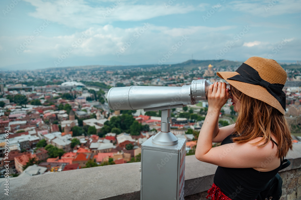 Woman in sun hat looking through binoculars to Tbilisi city, Georgia