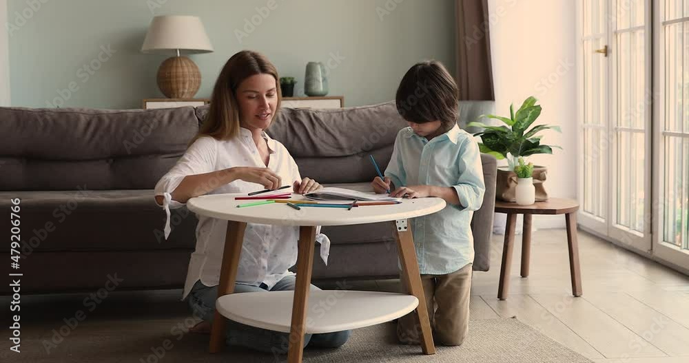 Happy young single mom little son friends sit on heated floor on carpet by small table draw funny pencil sketches together. Affectionate foster mum watch adopted kid boy preschooler creating picture Happy young single mom little son friends sit on heated floor on carpet by small table draw funny pencil sketches together. Affectionate foster mum watch adopted kid boy preschooler creating picture