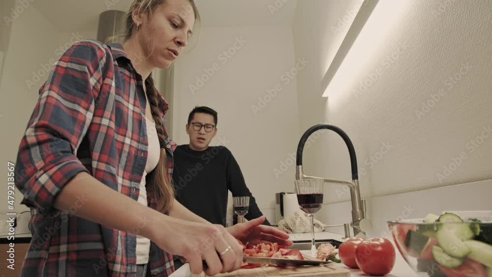 Woman talks to her asian boyfriend in the kitchen during chopping vegetables and cooking dinner. Couple cooking together in evening at home Woman talks to her asian boyfriend in the kitchen during chopping vegetables and cooking dinner. Couple cooking together in evening at home