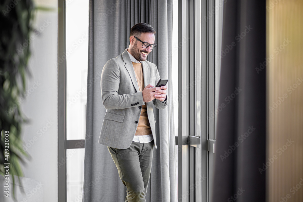 Smiling man in casuals standing in office. Businessman with mobile phone in hand. Smiling businessman using phone in office. Small business entrepreneur looking at his mobile phone and smiling.