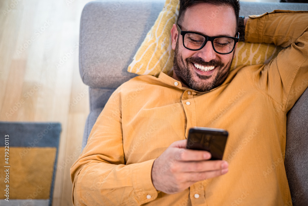 Cheerful young adult mature man happy smiling using smart phone resting lying down on sofa couch at home reading messages on mobile phone Guy with eyeglasses relaxing at home surfing net social media