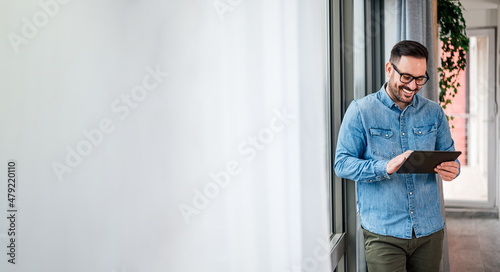 Smiling man businessman in casuals standing in office next to window using tablet making online shopping purchasing Small business home office entrepreneur looking at tablet making online video call