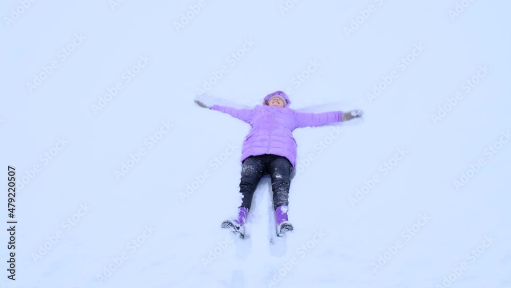 High angle girl wearing winter clothes color Very Peri, lying on back on white snow, making snow angel, moving hands to sides and back. High angle girl wearing winter clothes color Very Peri, lying on back on white snow, making snow angel, moving hands to sides and back.
