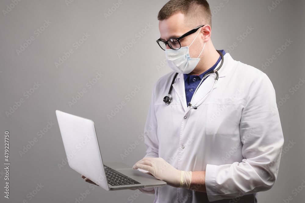 Successful young caucasian male doctor in a white medical uniform and with a stethoscope, busy working on a laptop, typing on a computer, consulting a patient online. Studio portrait Successful young caucasian male doctor in a white medical uniform and with a stethoscope, busy working on a laptop, typing on a computer, consulting a patient online. Studio portrait