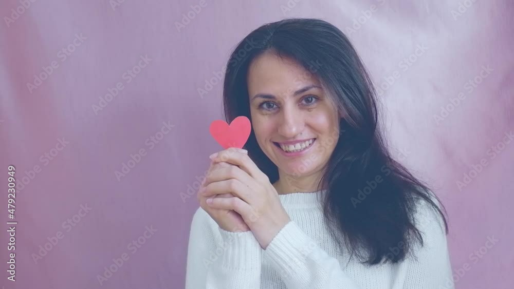 Smiling funny young brunette in a white sweater smiles, blinks and waving red hearts on a pink background. Valentine's Day