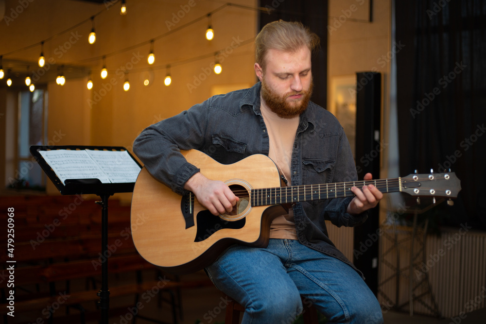 Obraz premium A young guy with a beard plays an acoustic guitar in a room with warm lighting