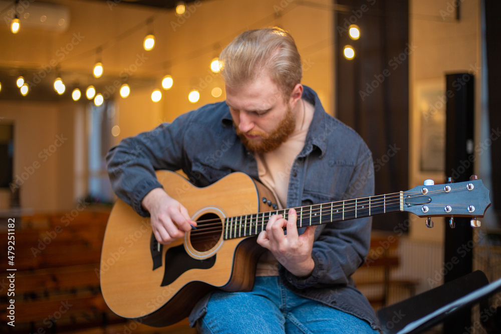 Obraz premium A young guy with a beard plays an acoustic guitar in a room with warm lighting