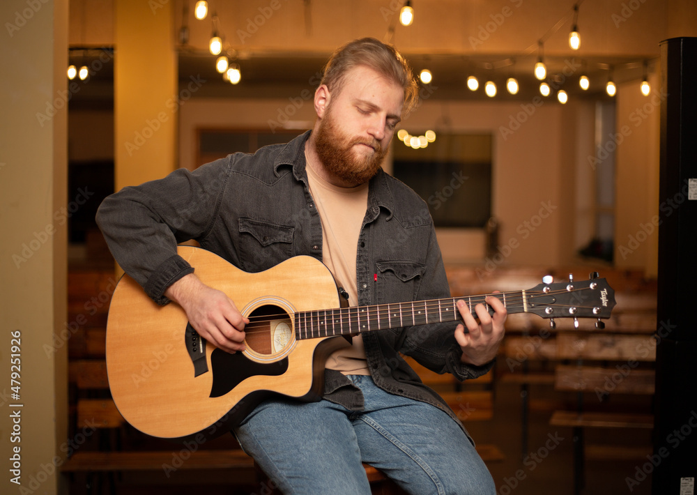 Obraz premium A young guy with a beard plays an acoustic guitar in a room with warm lighting