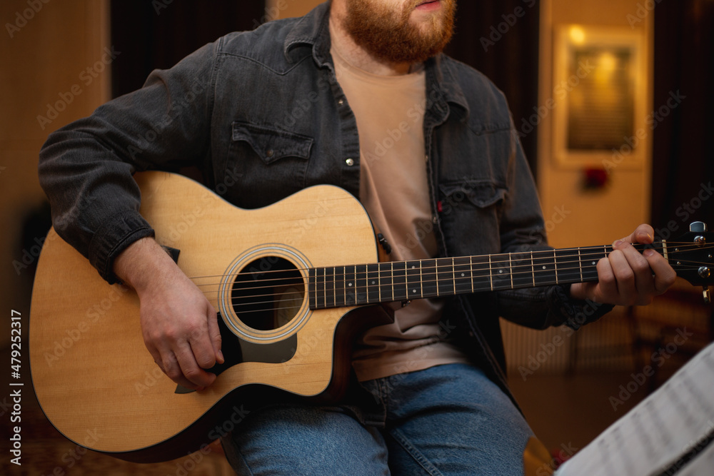 Obraz premium A young guy with a beard plays an acoustic guitar in a room with warm lighting