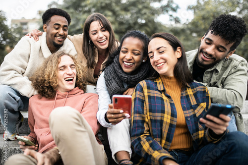 Happy group of young people having fun sharing video content on social media platform - Millennial multiracial friends using smartphone while sitting outdoors - Youth and technology concept