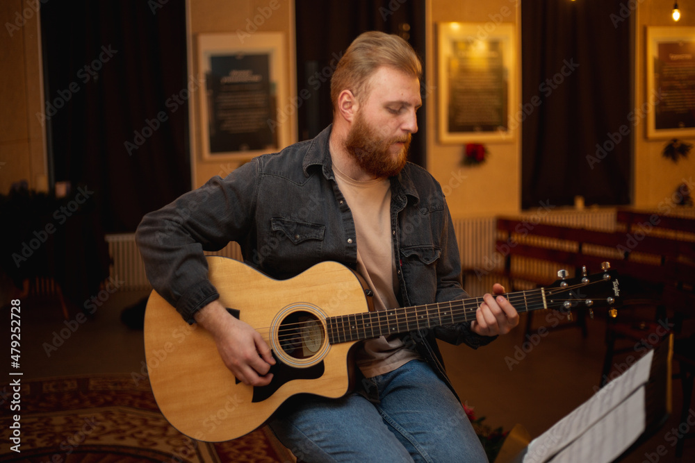 Obraz premium A young guy with a beard plays an acoustic guitar in a room with warm lighting