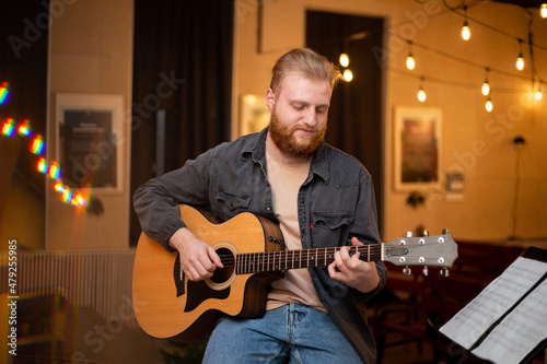 A young guy with a beard plays an acoustic guitar in a room with warm lighting
