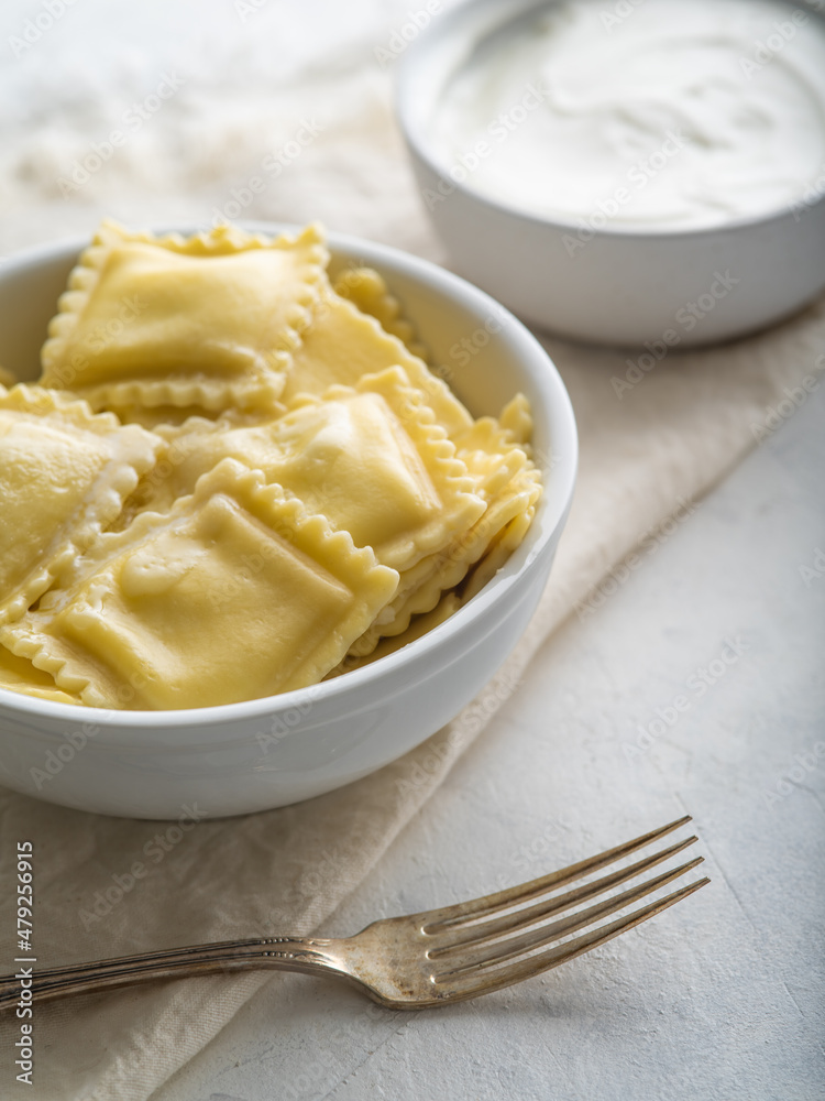Italian breakfast for one person. Ravioli with butter in a white earthenware bowl, fork and sour cream sauce. White tablecloth. Close-up. There are no people in the photo. Pastel shades. Italian breakfast for one person. Ravioli with butter in a white earthenware bowl, fork and sour cream sauce. White tablecloth. Close-up. There are no people in the photo. Pastel shades.