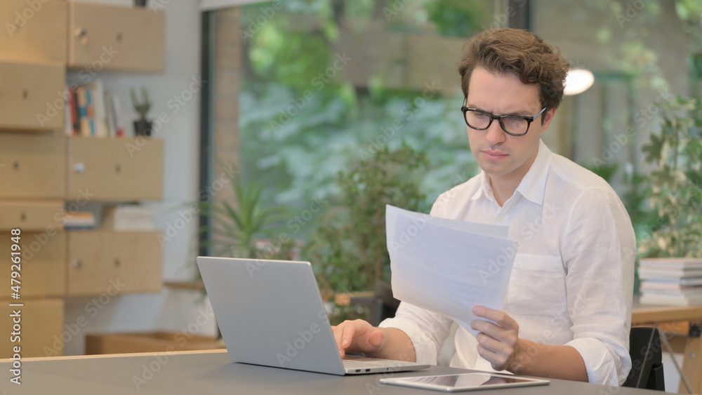 Man with Laptop Reading Documents in Modern Office