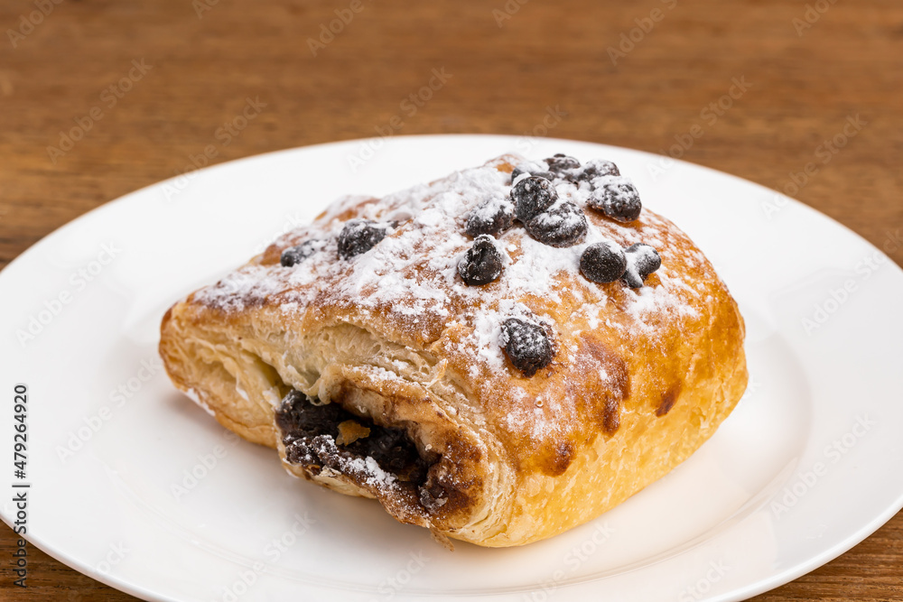 Single piece of Danish Pastry filled with chocolate cream topping with chocolate chips and icing sugar powder in white ceramic dish on wooden table.