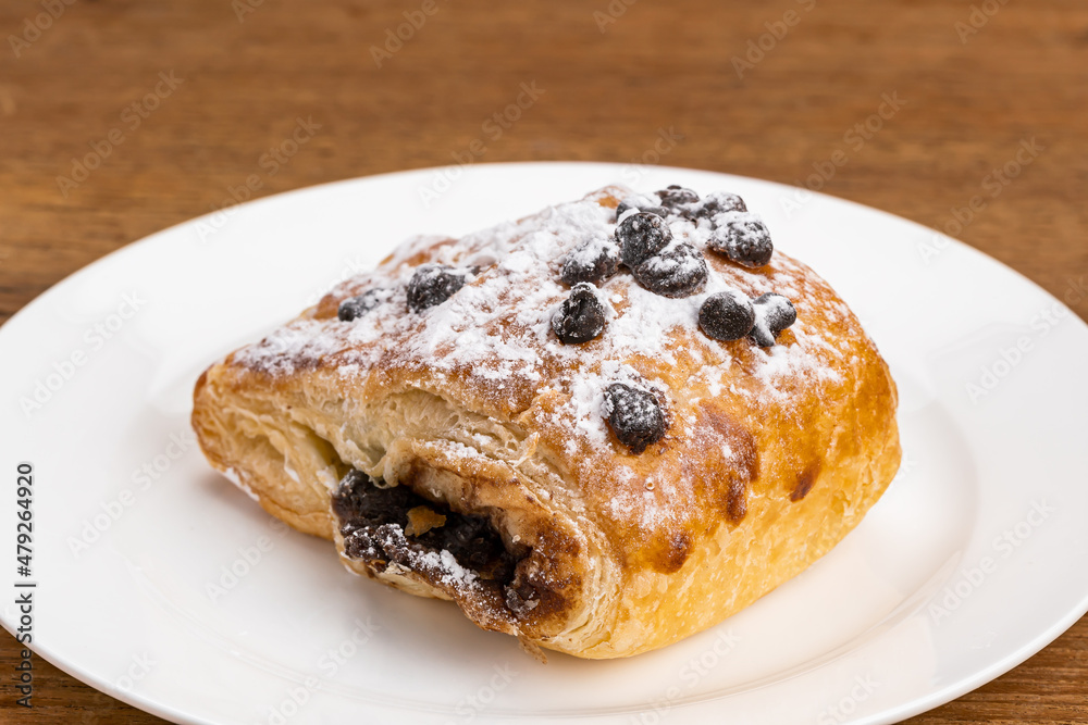 Single piece of Danish Pastry filled with chocolate cream topping with chocolate chips and icing sugar powder in white ceramic dish on wooden table. Single piece of Danish Pastry filled with chocolate cream topping with chocolate chips and icing sugar powder in white ceramic dish on wooden table.