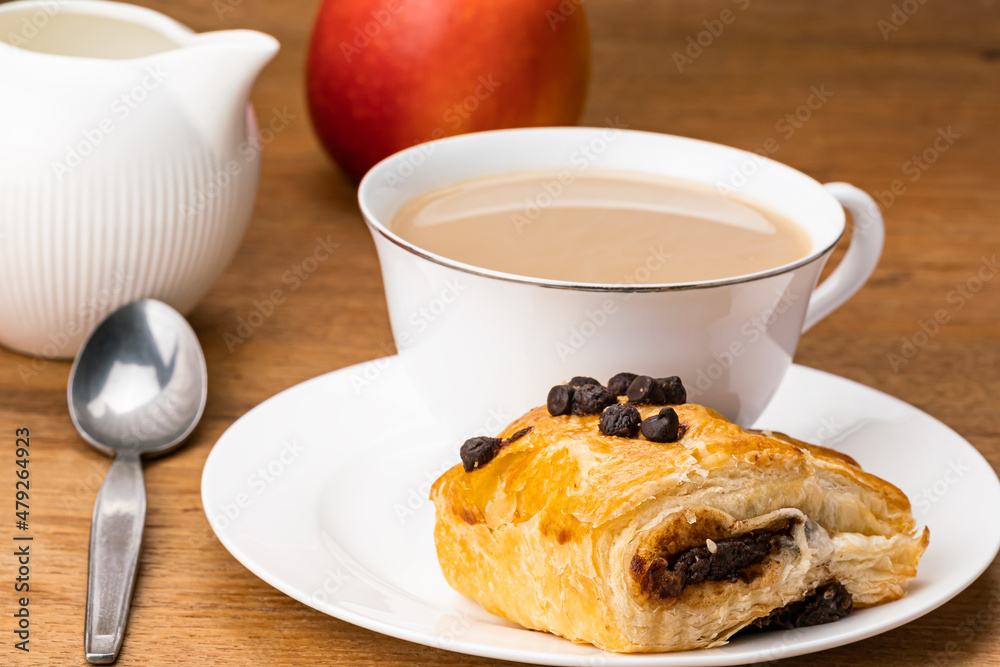 Danish Pasty filled with chocolate cream topping with chocolate chips with milk coffee in white ceramic cup on white saucer and ripe red apple on wooden table for breakfast. Danish Pasty filled with chocolate cream topping with chocolate chips with milk coffee in white ceramic cup on white saucer and ripe red apple on wooden table for breakfast.