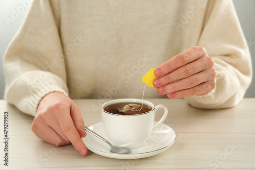 Woman dipping tea bag into cup of water at white wooden table, closeup