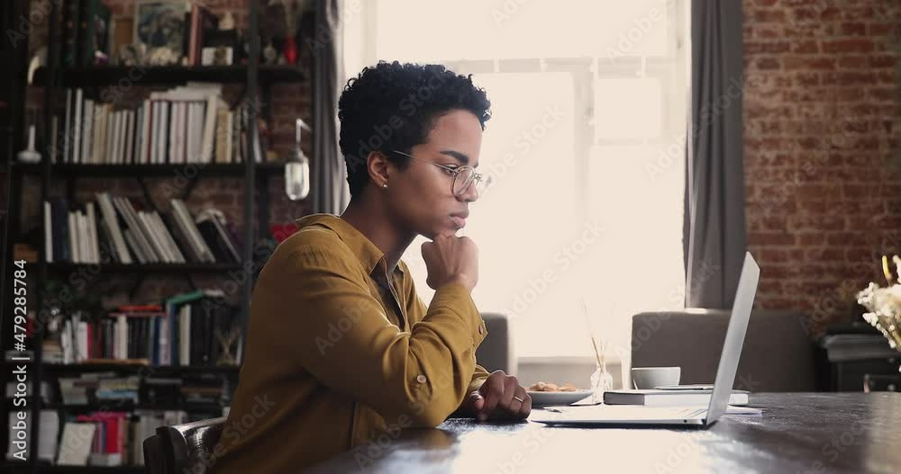 Serious African woman sit at desk working on laptop hold document, do paperwork looks thoughtful. Workflow, modern tech, online project, student prepare for exams makes assignment use computer concept