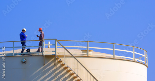 Engineer and worker are working to check oil quality on top of big fuel storage tank against blue clear sky background